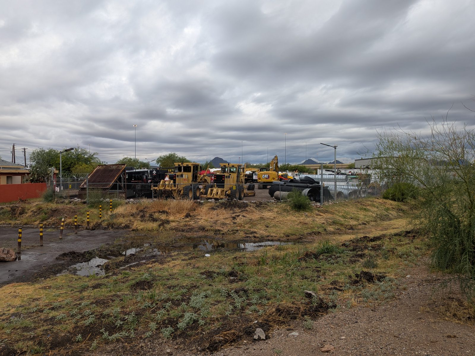 View north across the alley showing standing water in the regulatory wash, Magnum Paving heavy equipment behind the chain-link fence, and stacked drainage culverts.